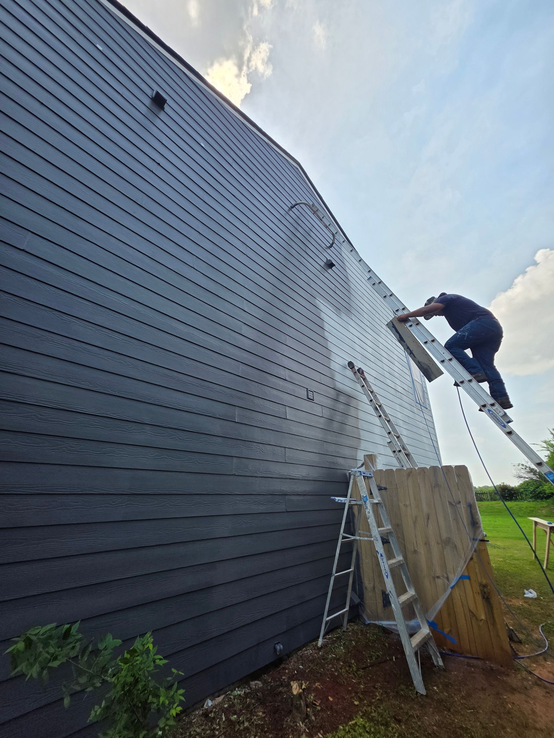 Man on a ladder installing siding on a dark gray building exterior, sunny day.