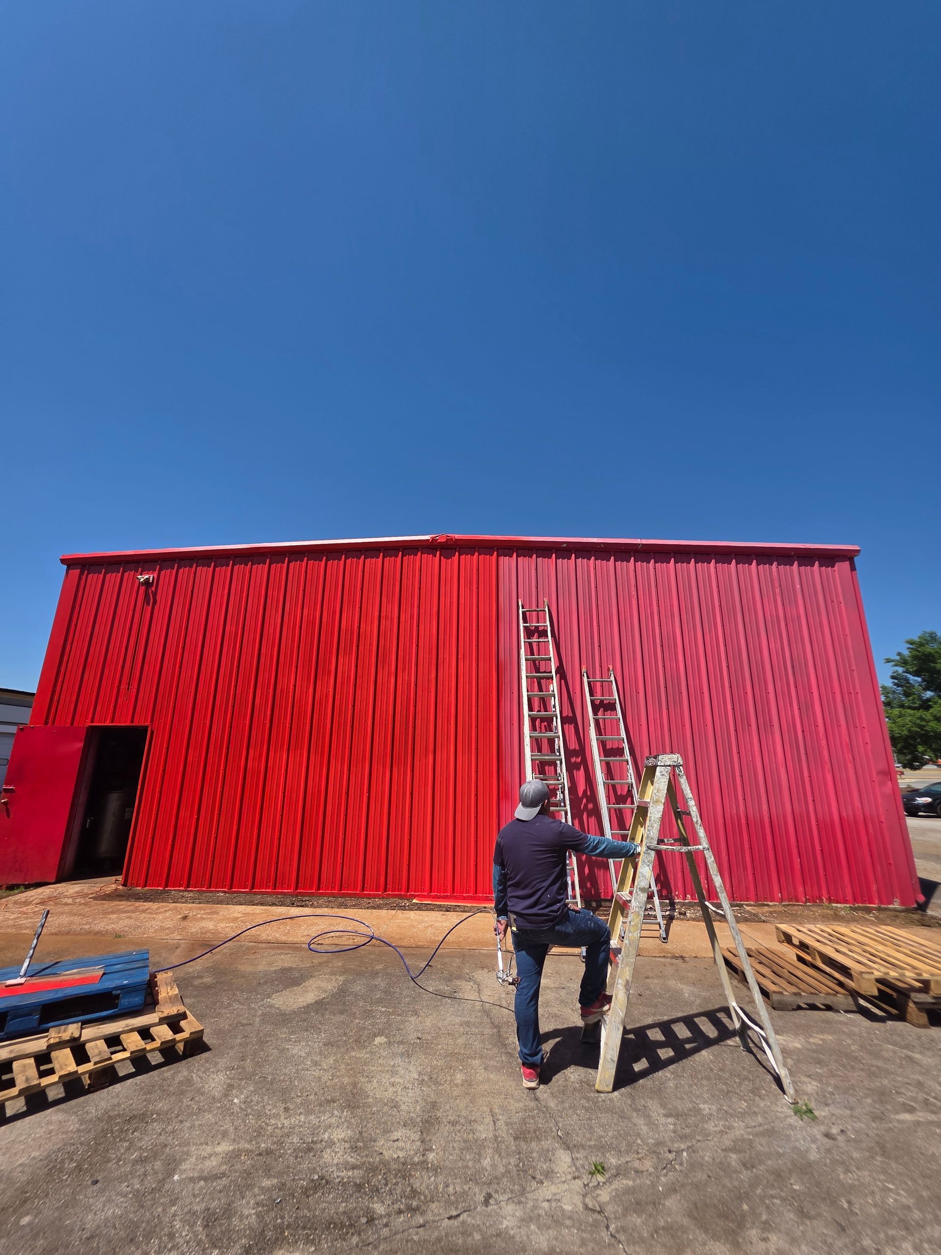 Person painting a red building, standing on a ladder. The sky is blue.