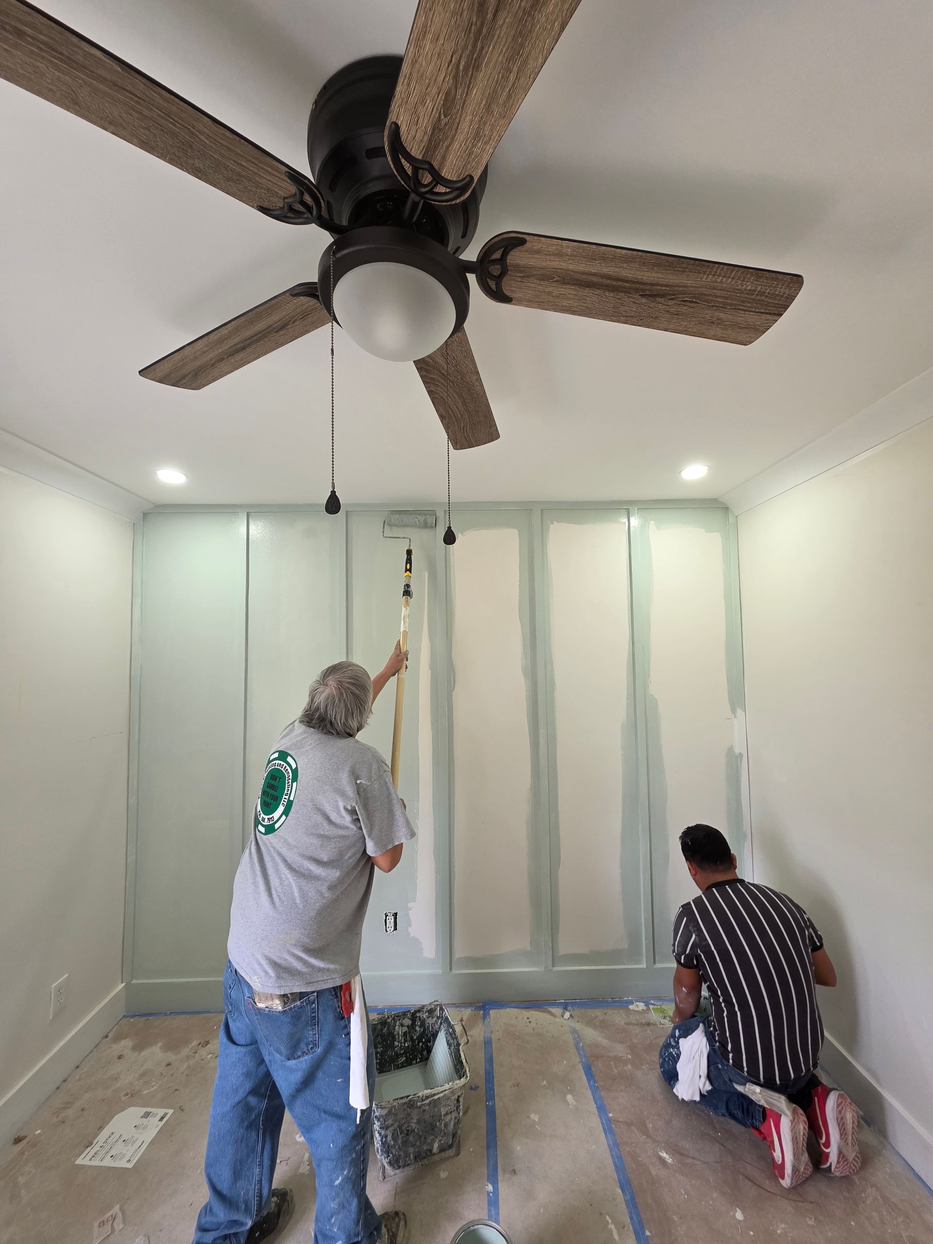 Two people painting a room. One standing, the other kneeling. A ceiling fan hangs above.