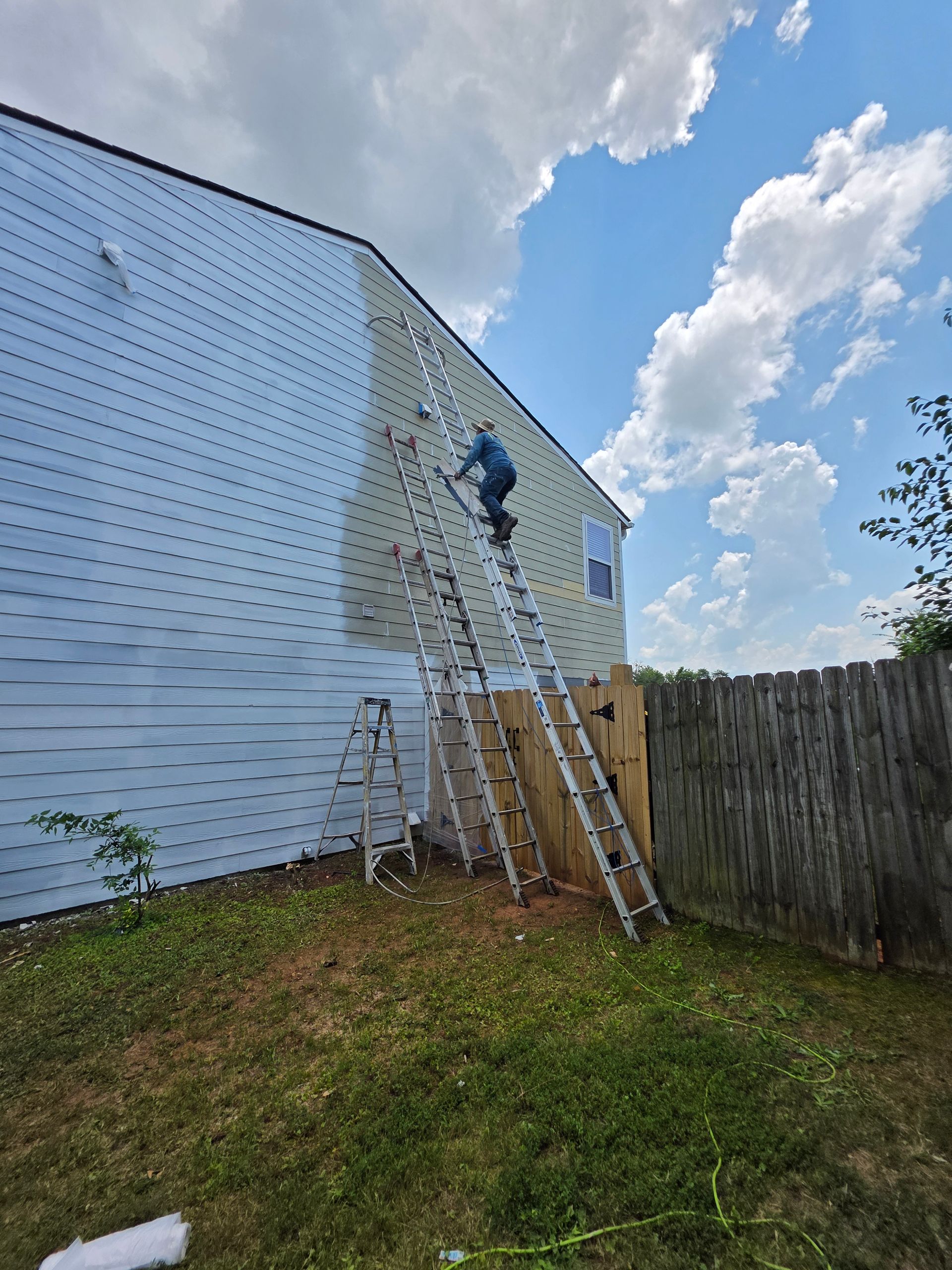 Person on ladder siding a house, blue sky, green grass, wooden fence.