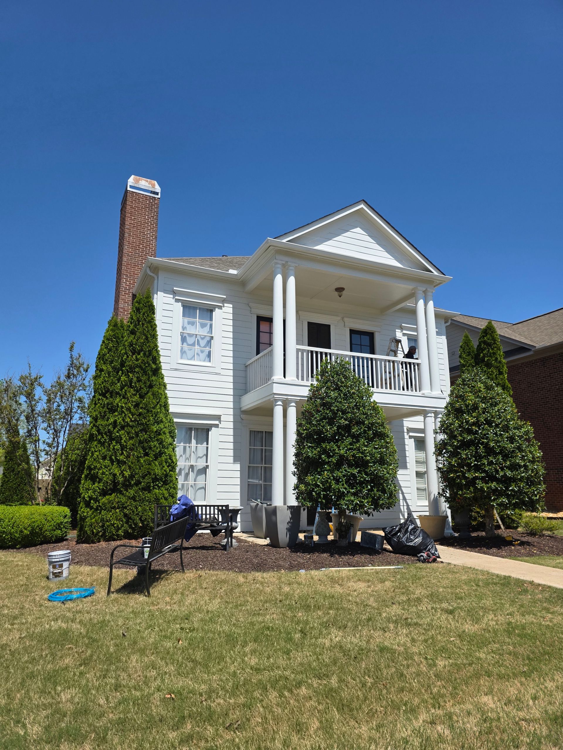 White two-story house with a porch, chimney, and trees on a sunny day.