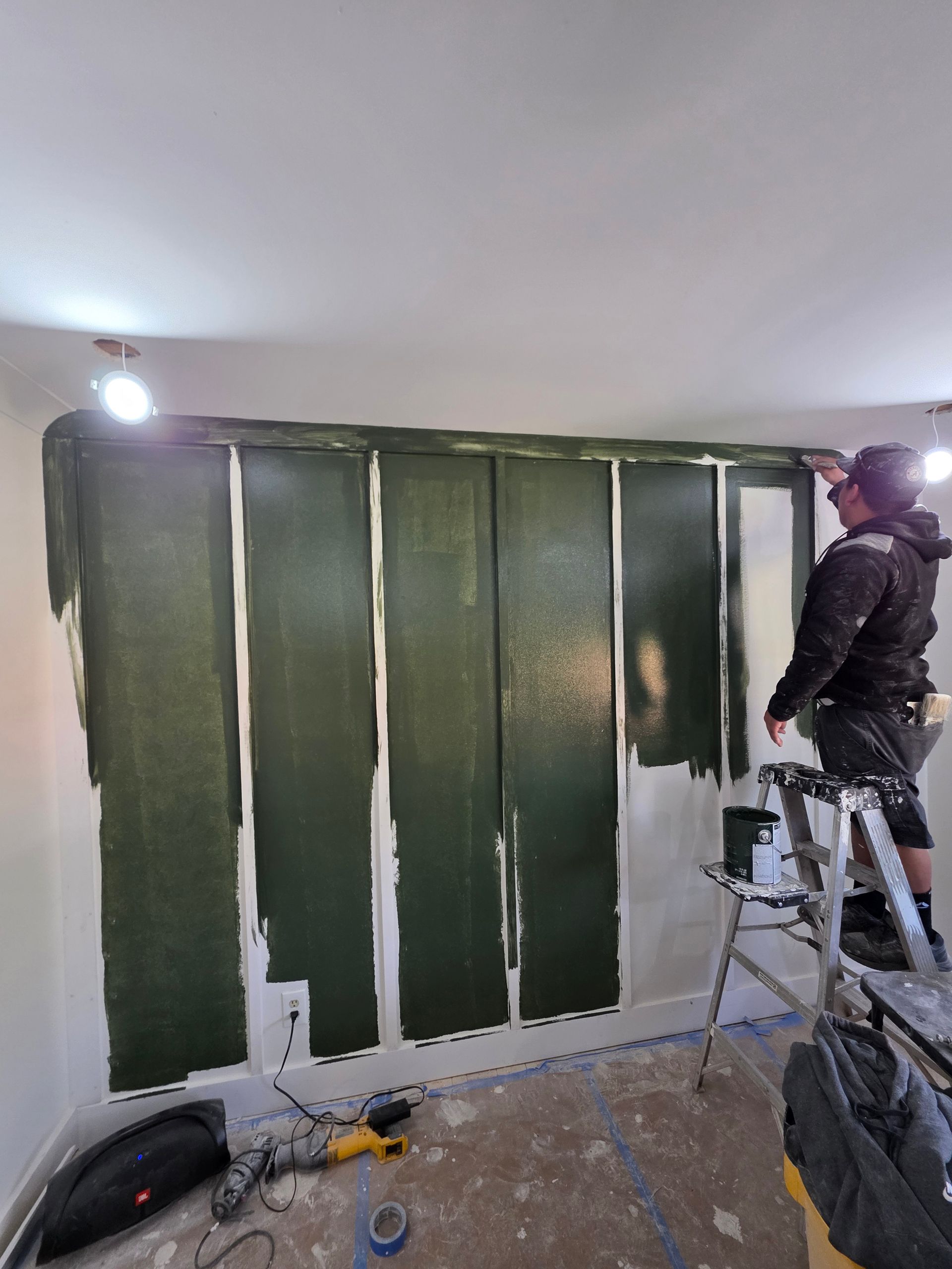 Person painting green stripes on a white wall. Ladder, paint supplies in room.