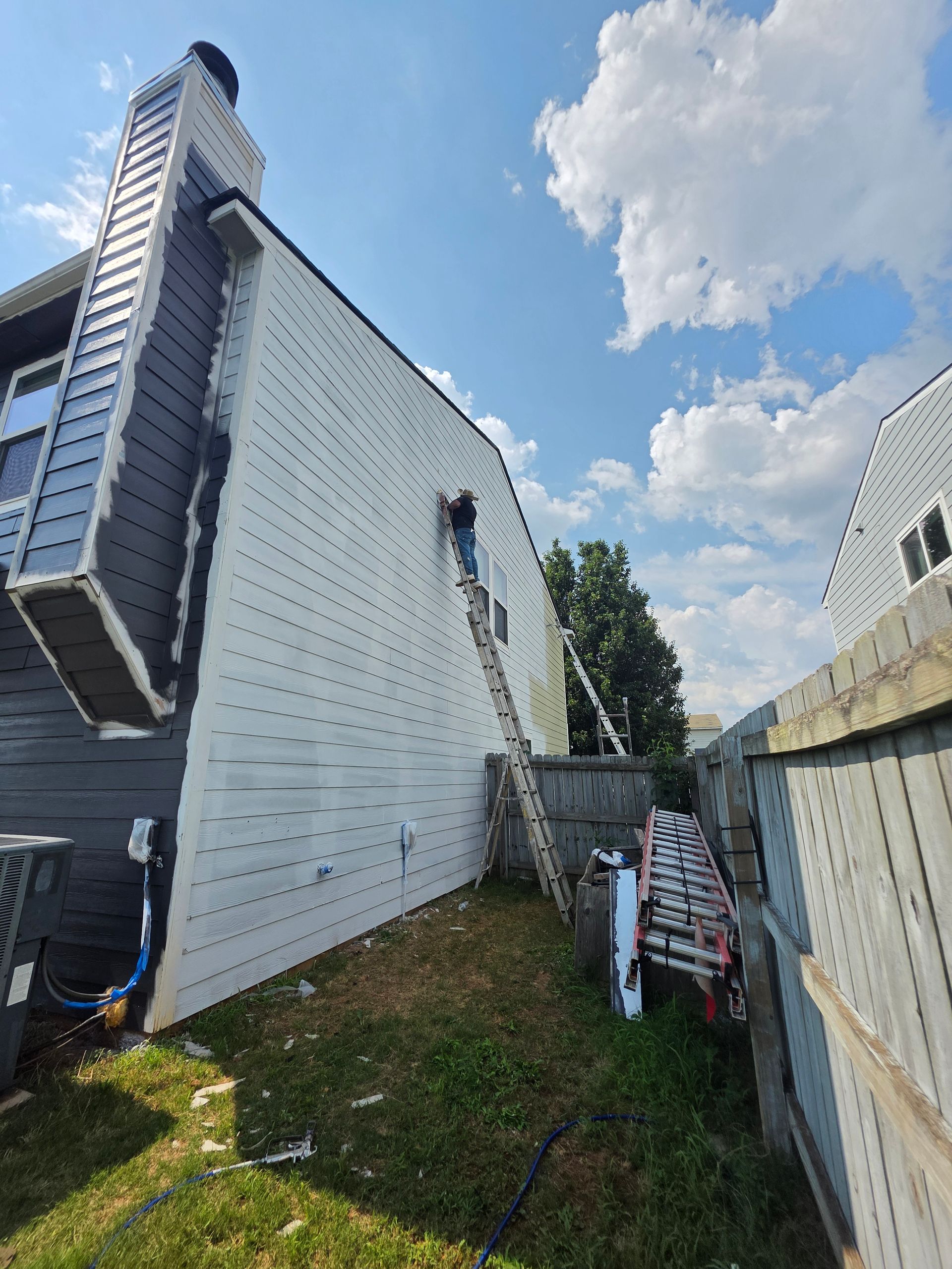 Exterior of a building with white siding and a chimney. A person on a ladder near the roof, working on the siding.