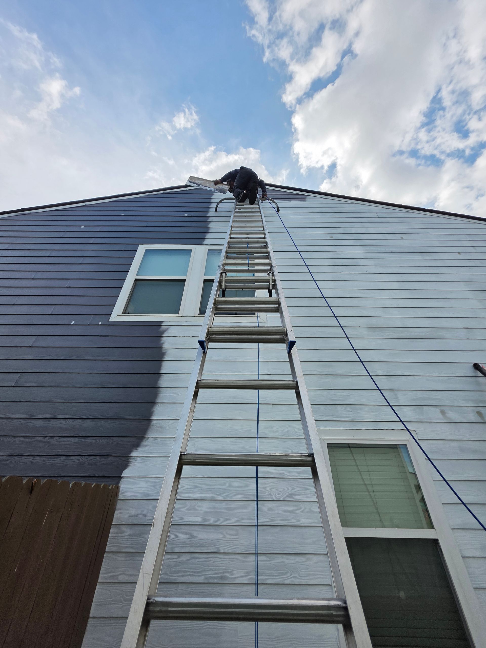 Person on ladder painting house blue and white against a cloudy sky.