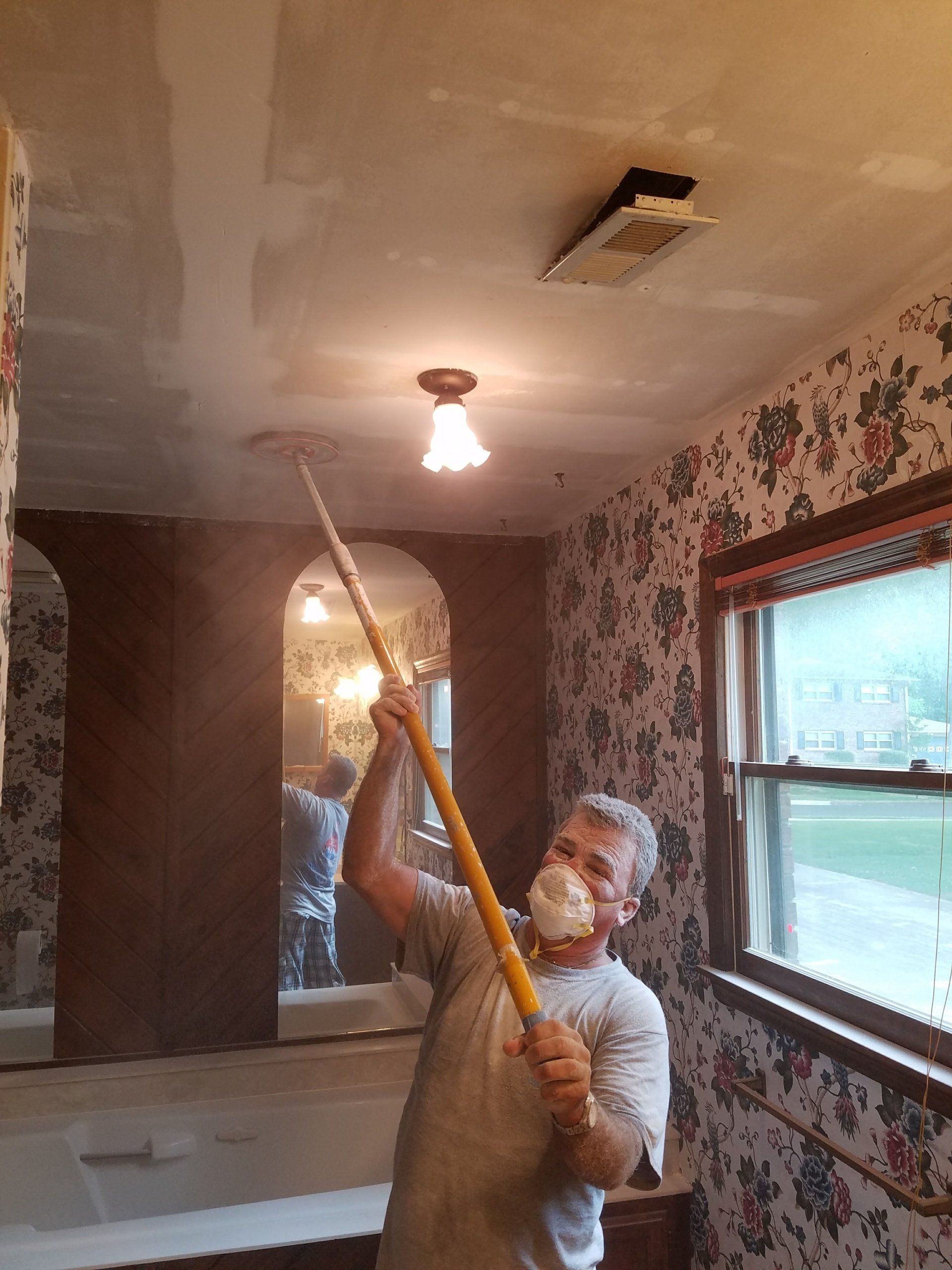 Man in a mask scraping a textured ceiling in a room with floral wallpaper. Another person in the background.