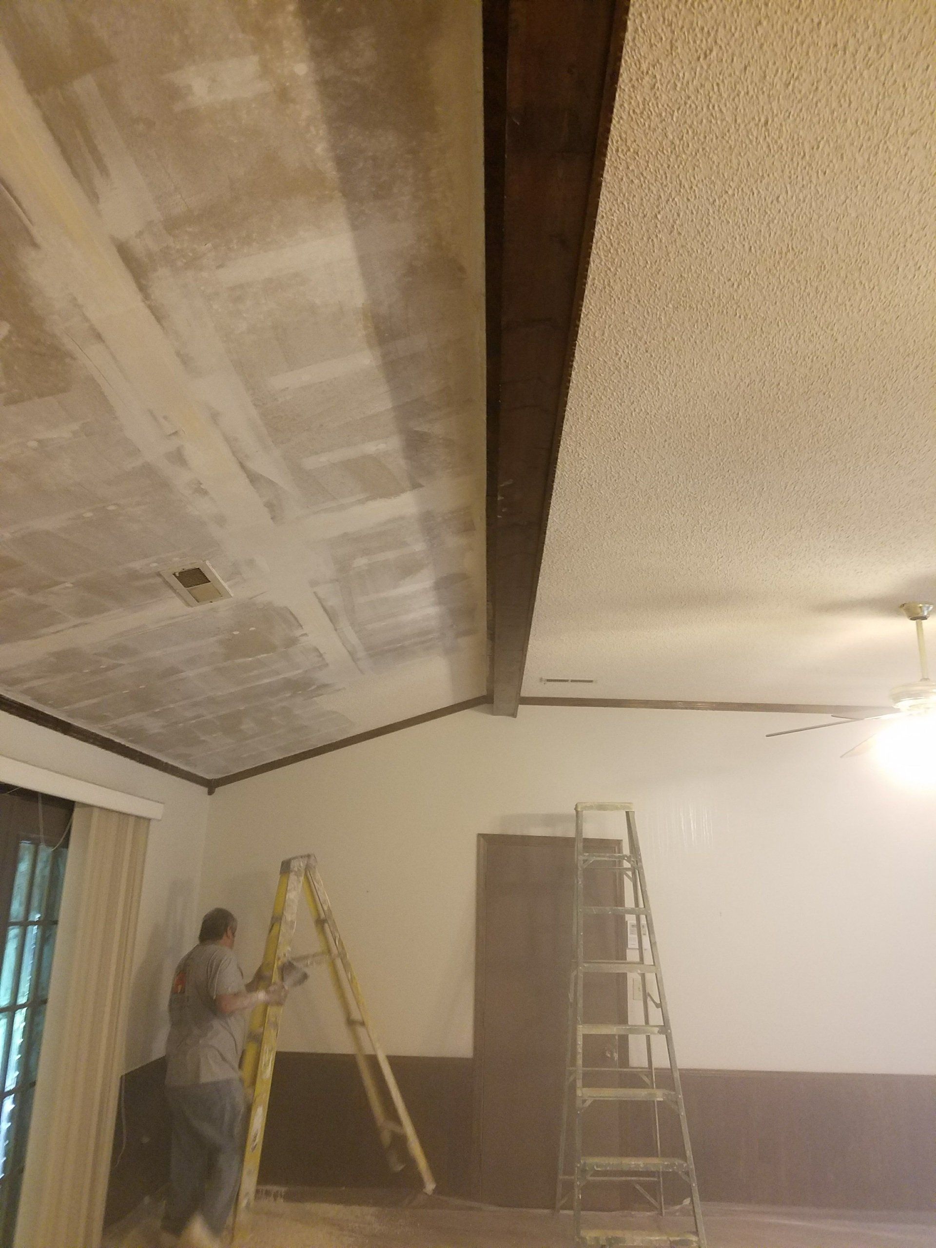 Man sanding ceiling, ladder beside him. Brown beam, white walls, and textured ceiling.