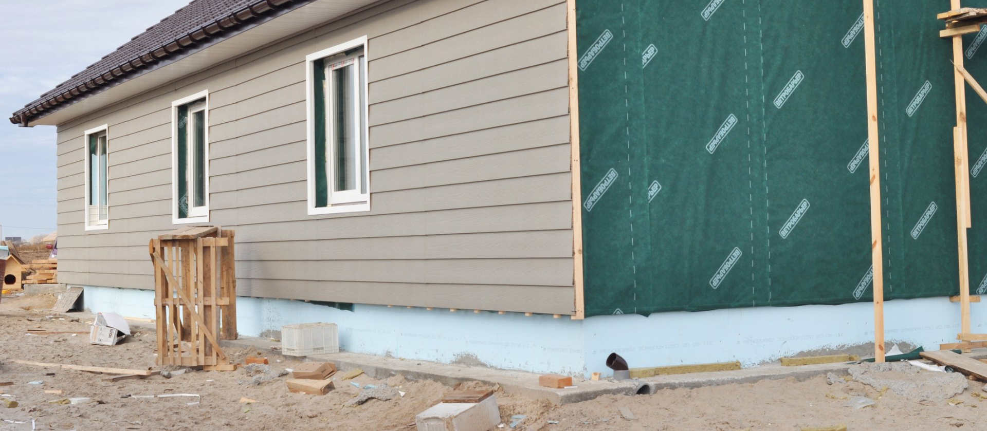 A house under construction; tan siding, windows, and exposed green wrap. Scaffolding and dirt visible.