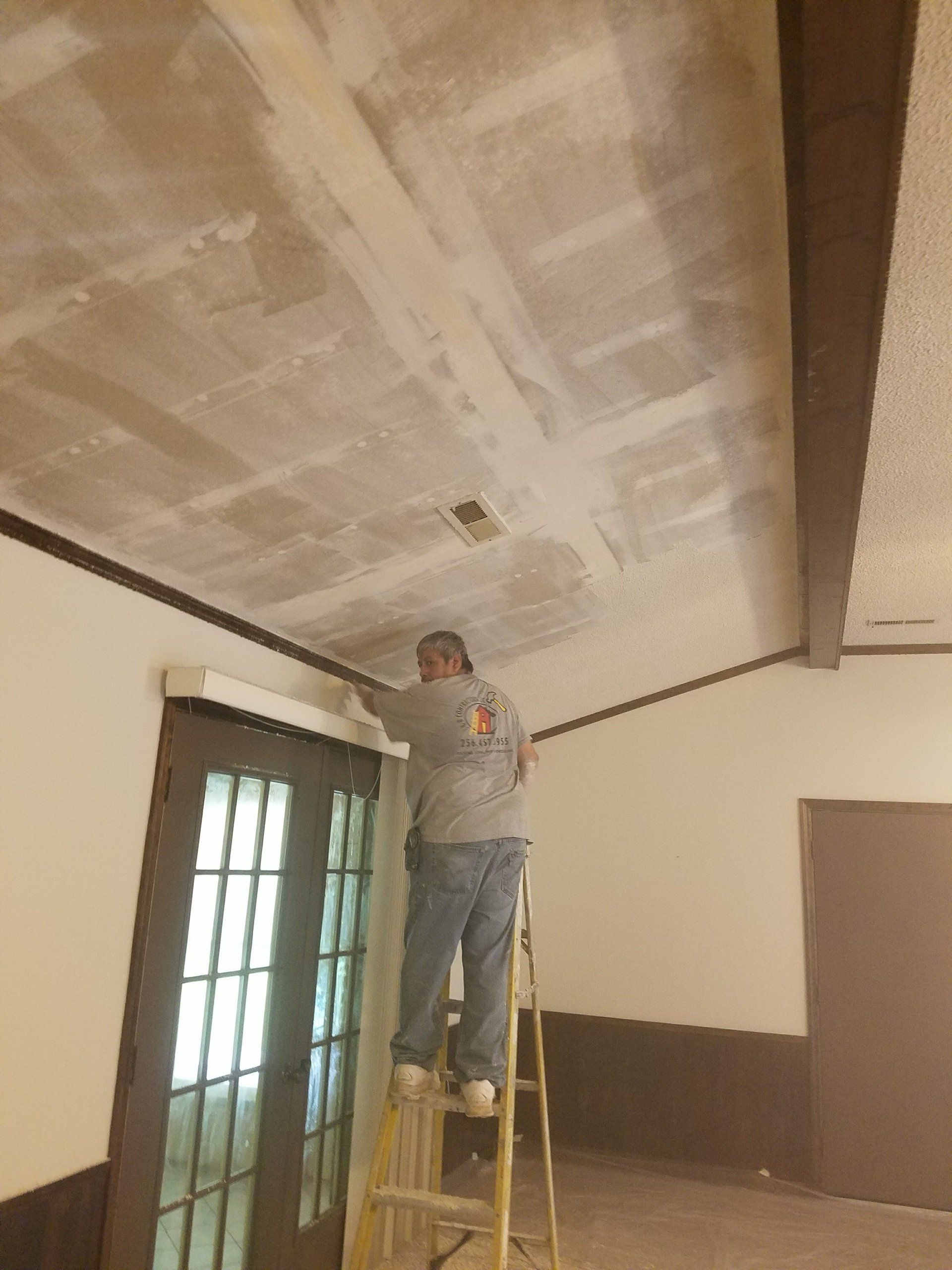 Man on a ladder, painting a ceiling with exposed beams and a door in the background.