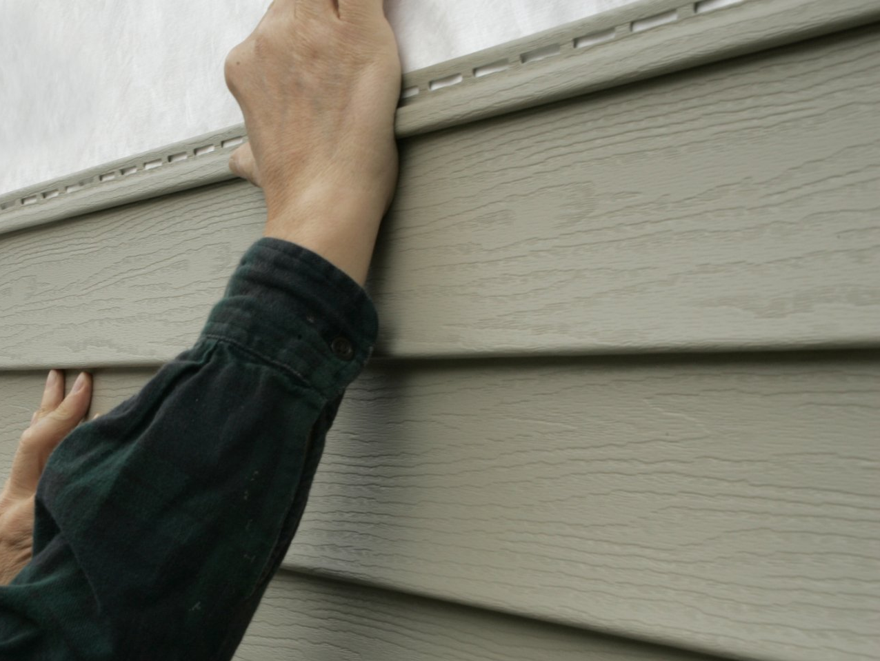Hands installing light brown vinyl siding on a building.