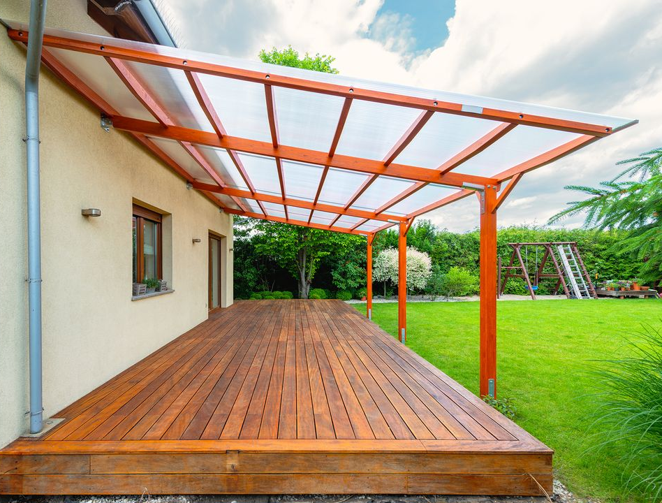 Wooden deck with a pergola, beside a house, surrounded by grass.