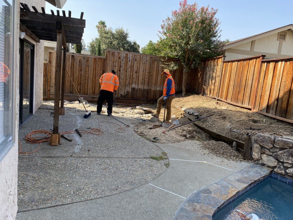 Two men are working on a patio in front of a wooden fence.