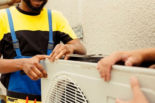A person in a yellow and black shirt and blue overalls works on an outdoor air conditioning unit.