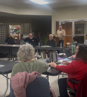 People at tables in meeting, one man speaks from a podium. Indoors, neutral colors.