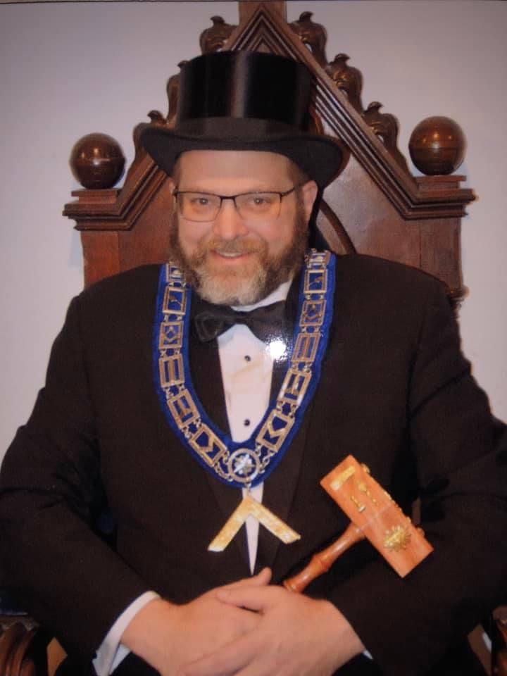 Man in suit and top hat seated, holding a gavel and wearing a Masonic collar in a wood-paneled room.