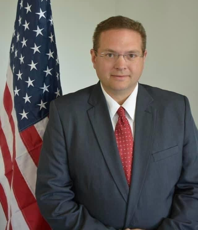 Man in suit and tie stands near an American flag.