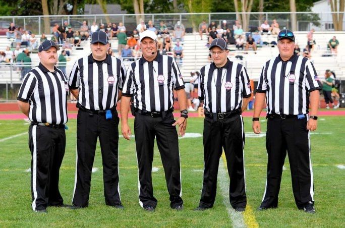 Five football referees in black and white striped shirts and black pants stand on a green field.