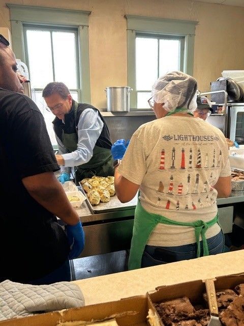 People preparing food in a kitchen. One wears a hairnet and apron. Others wear gloves and work at a counter.