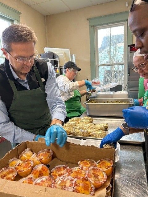 Volunteers preparing food in a kitchen. Man in apron looks at rolls, others portion food into containers.