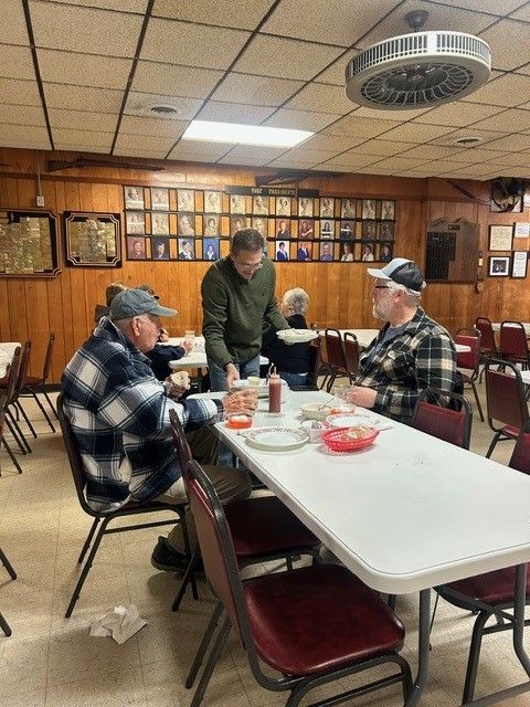 Three men at a table in a diner. One man stands serving the others food. Brown paneling and many photos on the wall.