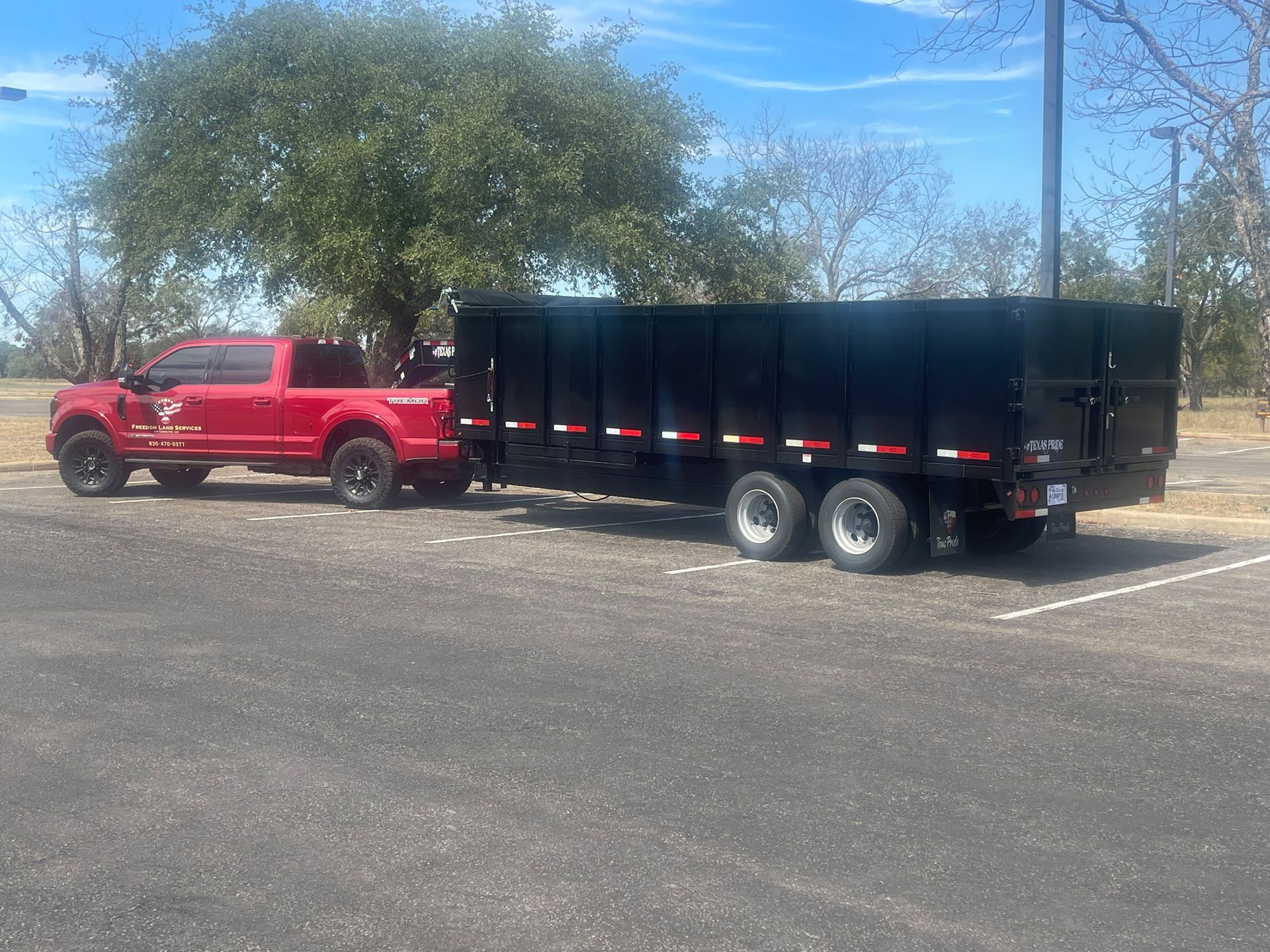 A red truck is towing a black dumpster in a parking lot.