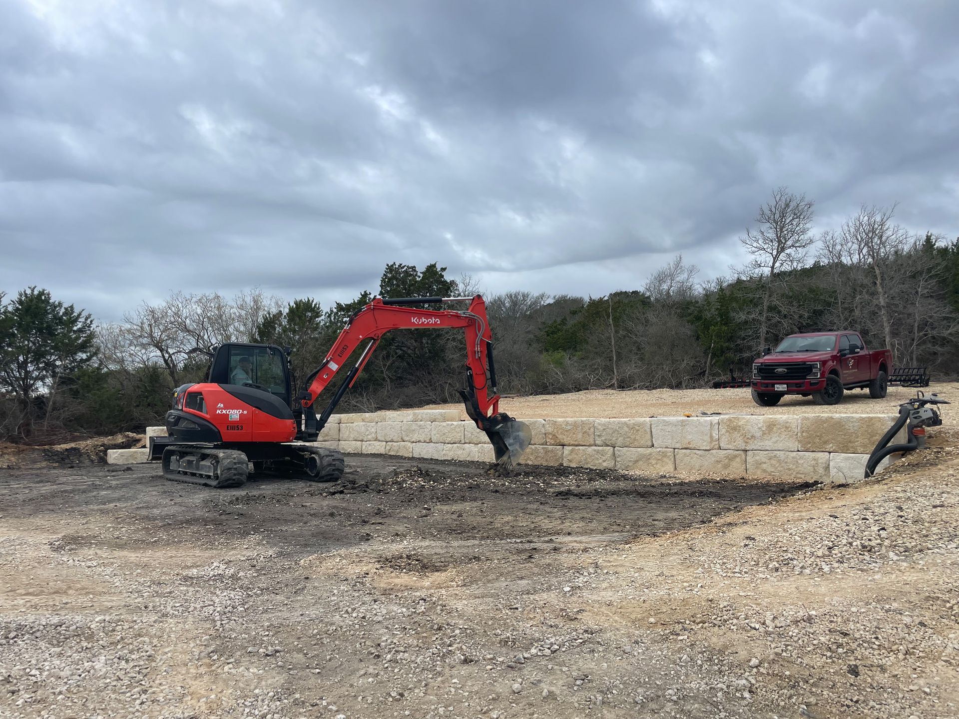 A red excavator is sitting in a gravel lot next to a red truck.