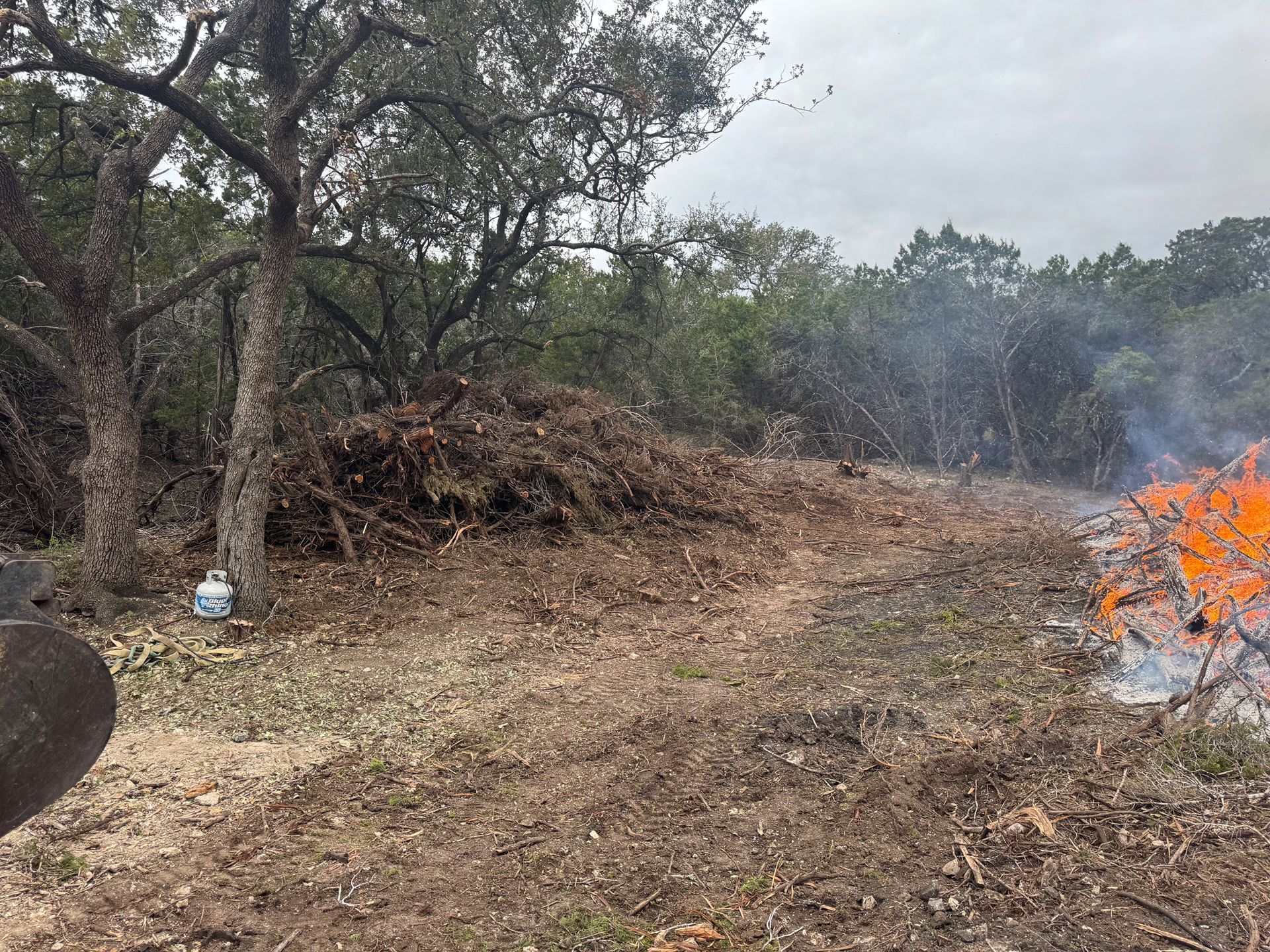 Pile of brush burning outdoors, producing smoke and flames in a wooded area.