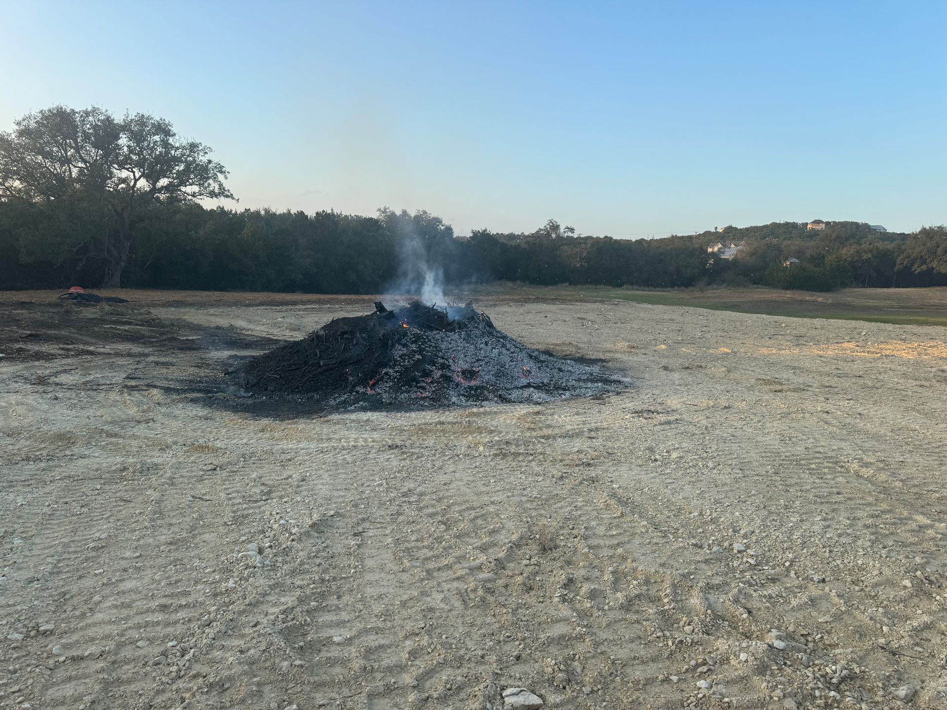 Burning pile of dark material in a clearing, emitting smoke. Trees in the background under a blue sky.