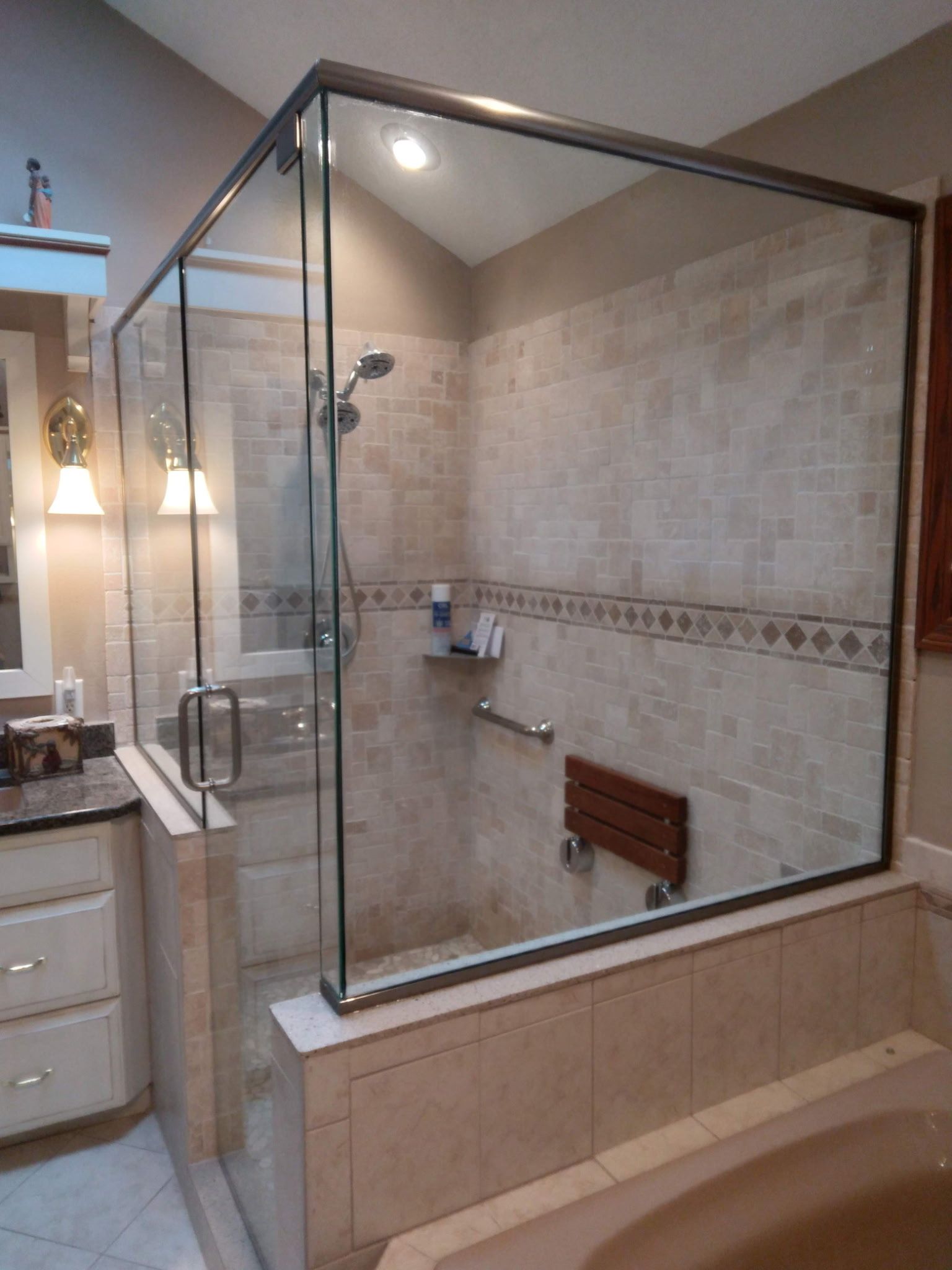 Glass shower enclosure in a bathroom with beige tile and a wooden seat.