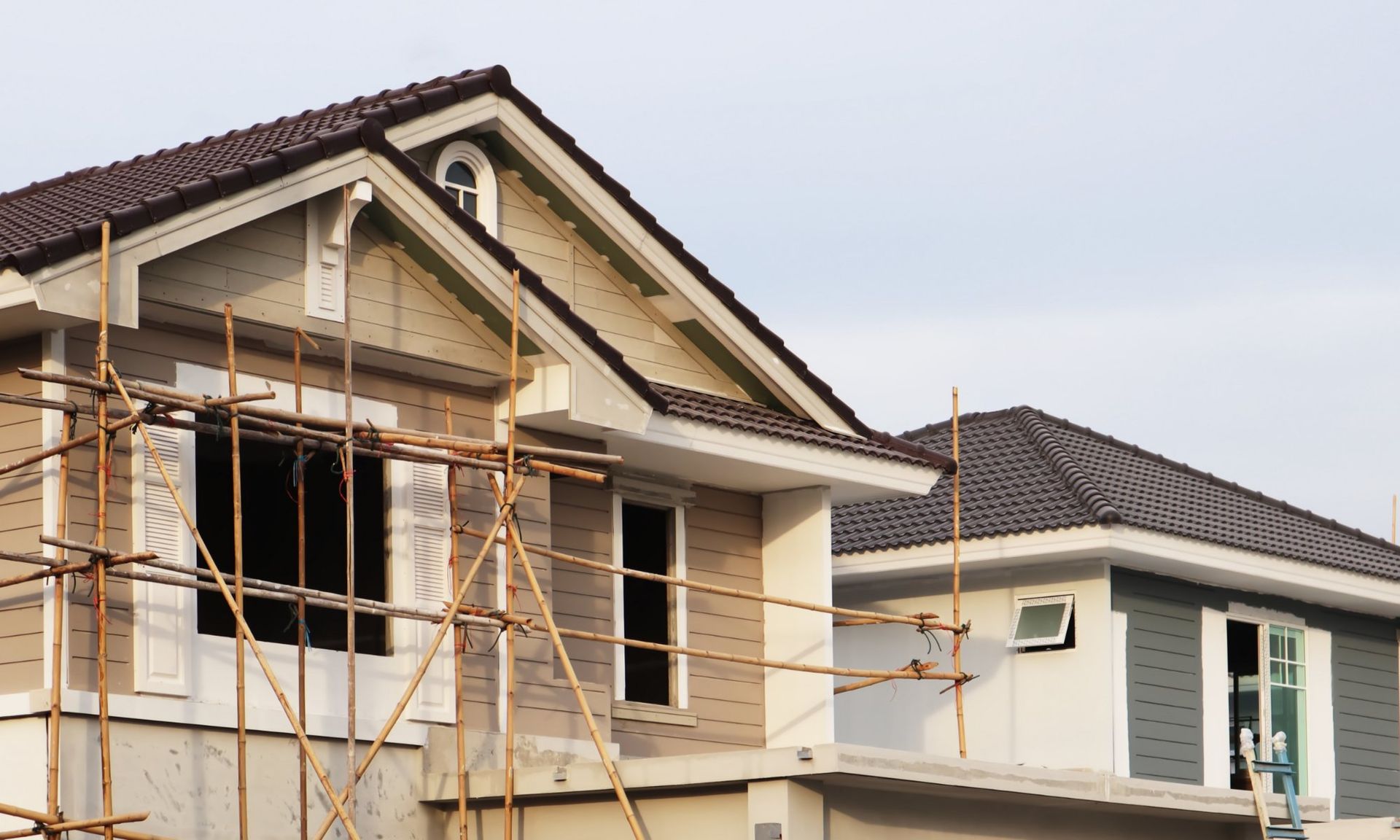 Two-story house under construction with scaffolding, beige and grey siding, brown roof, cloudy sky.