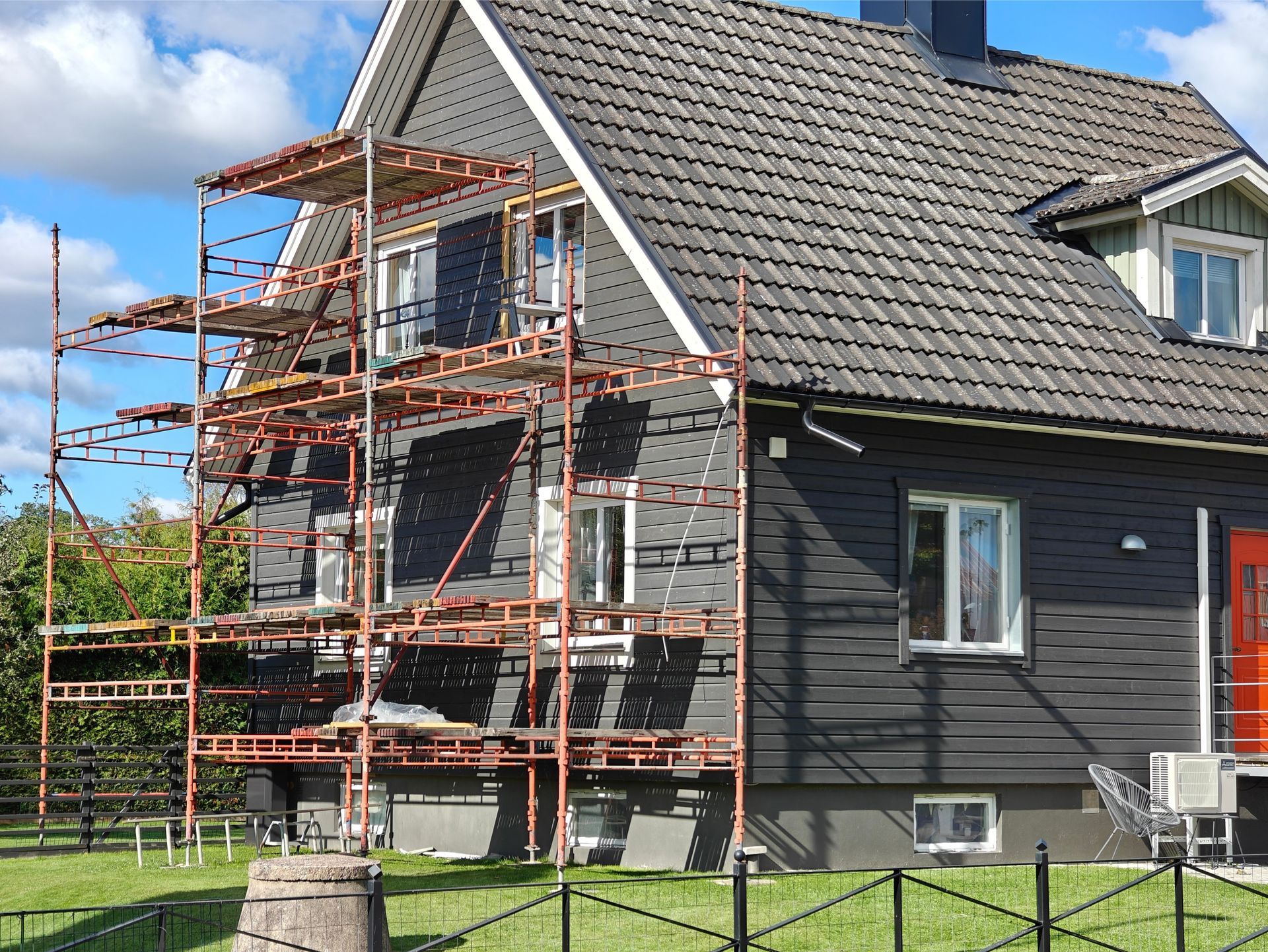 Scaffolding on a dark gray house with white window frames; lawn, blue sky, and a red door are visible.