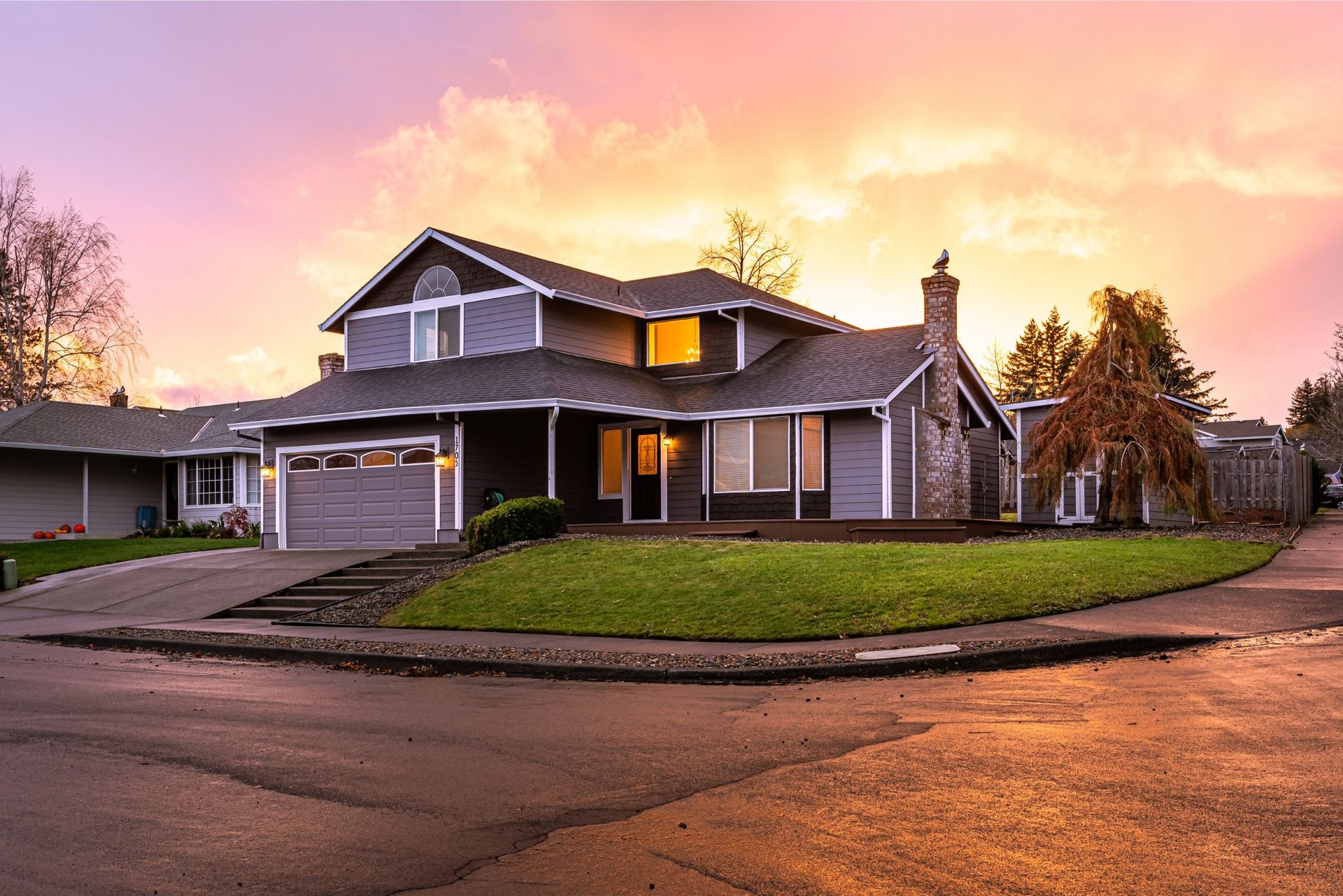 Two-story gray house at sunset. Features a garage, chimney, and green lawn.