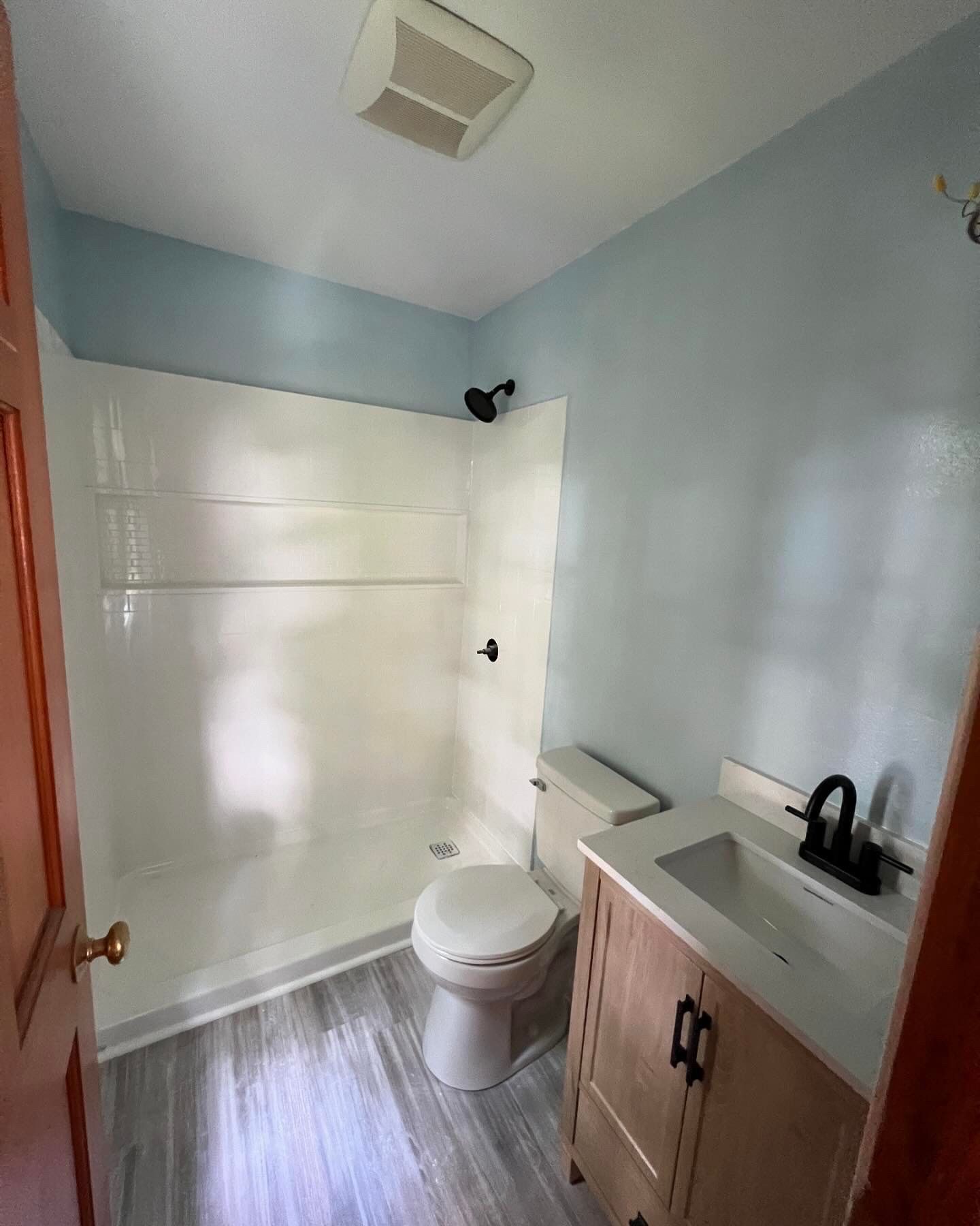Small bathroom with a white shower, toilet, and vanity. Light blue walls, wood door and gray floor.