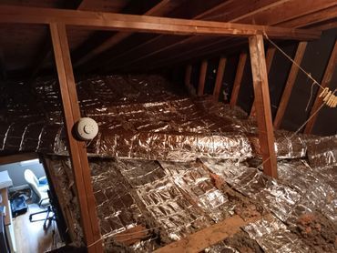 Attic space with reflective insulation and wooden framing. A smoke detector is visible, and a desk is seen below.