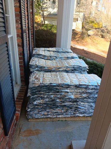 A porch stacked with numerous reflective insulation panels, next to a brick wall and window with shutters.