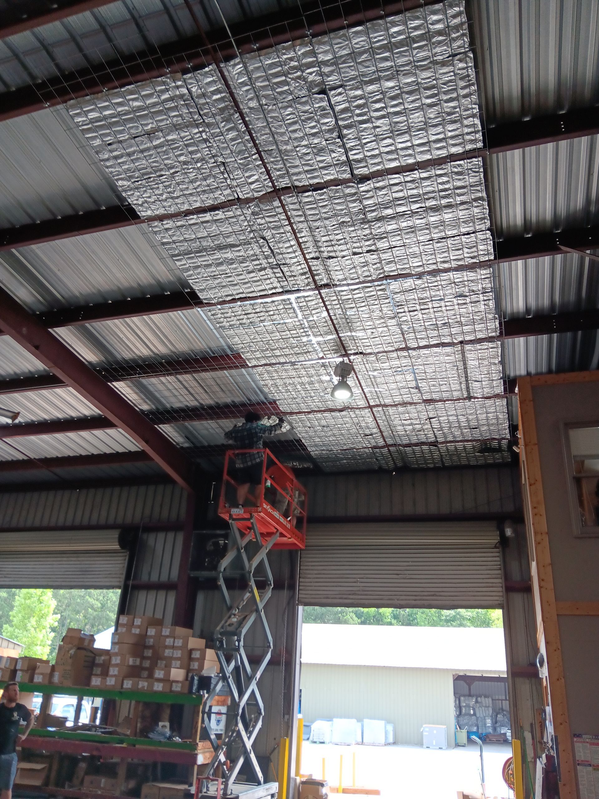 Warehouse interior with silver insulation panels installed on the ceiling, scissor lift visible, and a loading bay door.