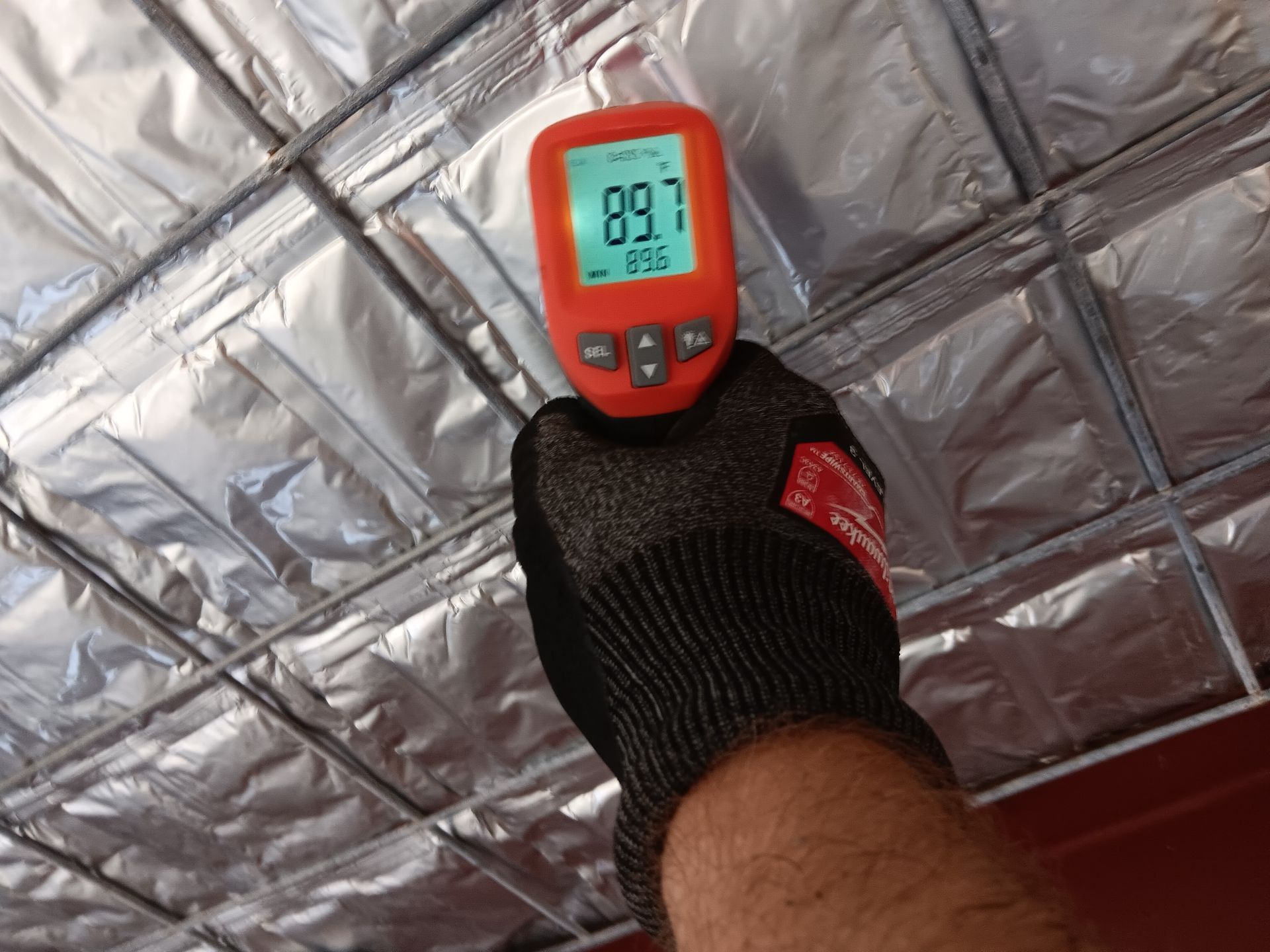 A gloved hand holds a digital thermometer measuring 89.7°F against silver insulation on a ceiling.