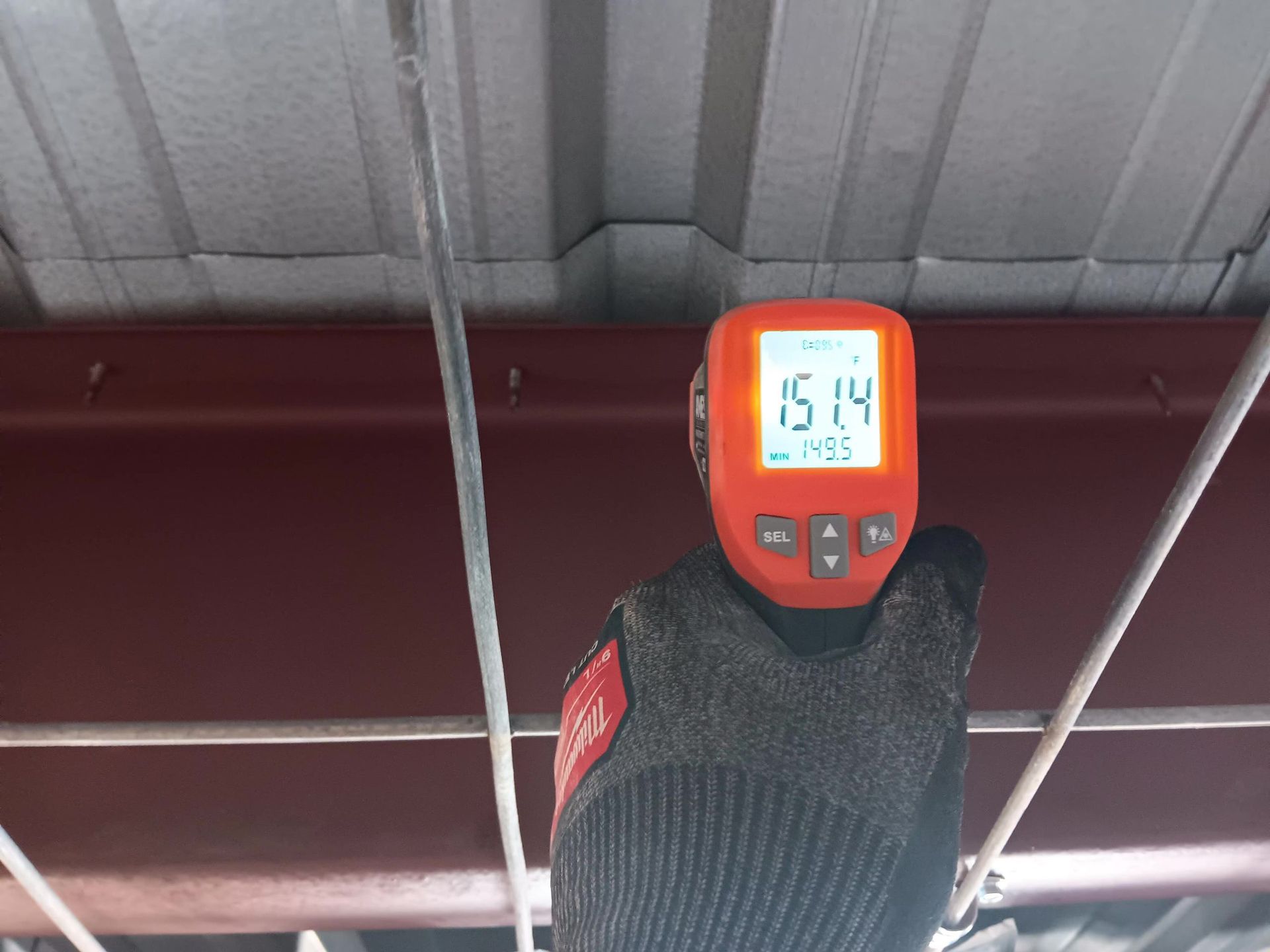 A person in a work glove holds a temperature reader to the metal roof's underside. Display reads 15.14°C.
