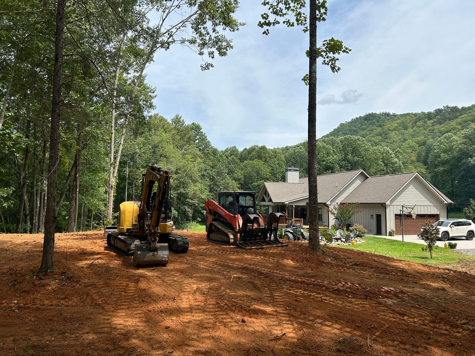 Excavators working on a red dirt lot beside a house in a wooded rural area