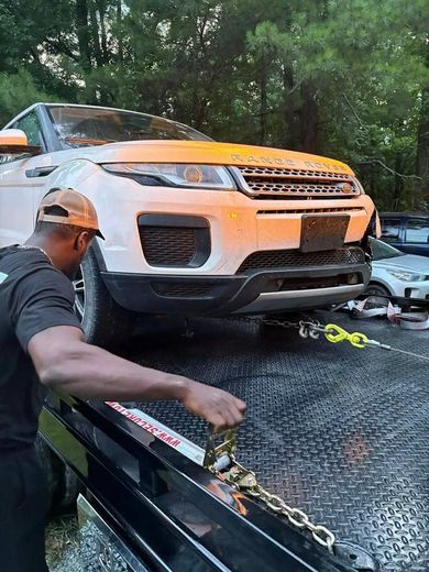 Man securing a damaged orange Range Rover on a tow truck. Forest setting.