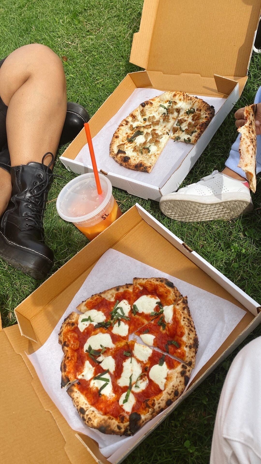 A person is sitting on the grass with boxes of pizza and a drink.