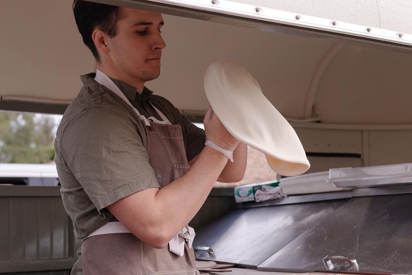 A man in an apron is holding a pizza dough in his hands, Beyond Basil Mobile Pizzeria.