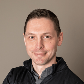 A man wearing a black jacket and a grey shirt is smiling for the camera.