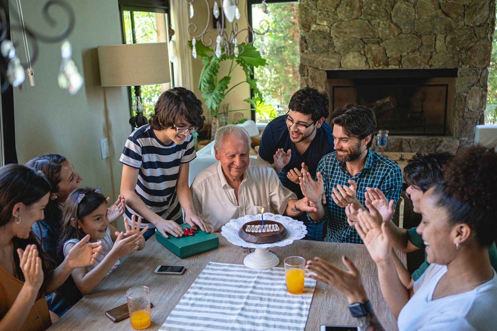 A family celebrates an elderly man's birthday, clapping as he smiles at a chocolate cake with candle