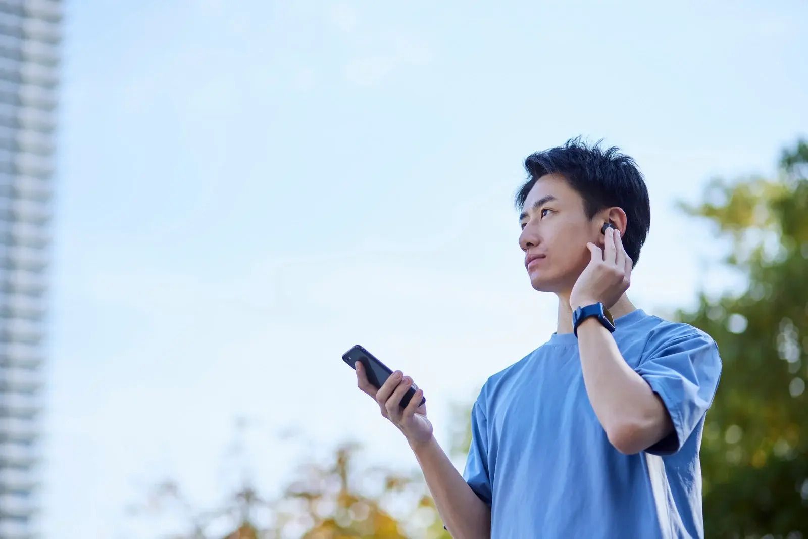 Person outdoors, wearing earbuds, holding phone, blue shirt, looking upward.
