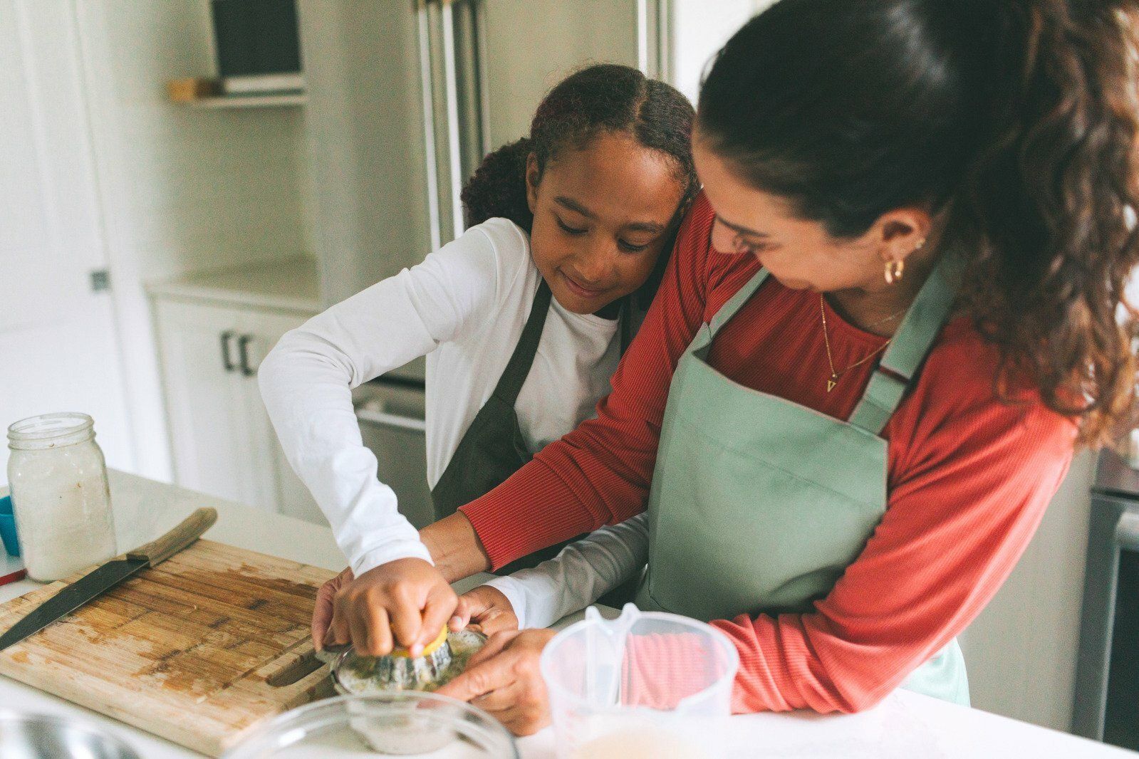 A woman and girl in aprons making dough in a bright kitchen.