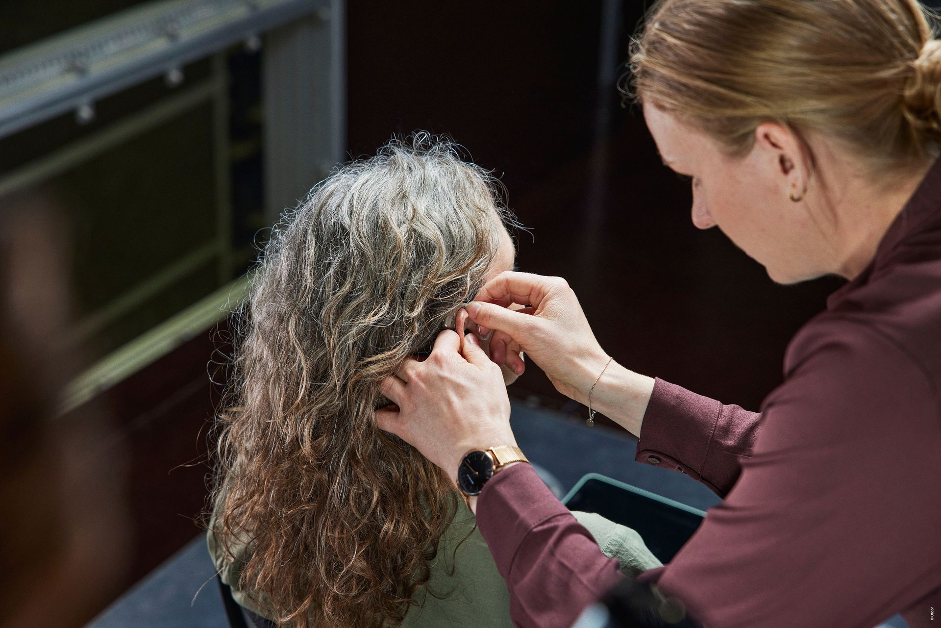 Audiologist fitting a hearing aid for a patient with curly gray hair at Audiology First, Lethbridge.