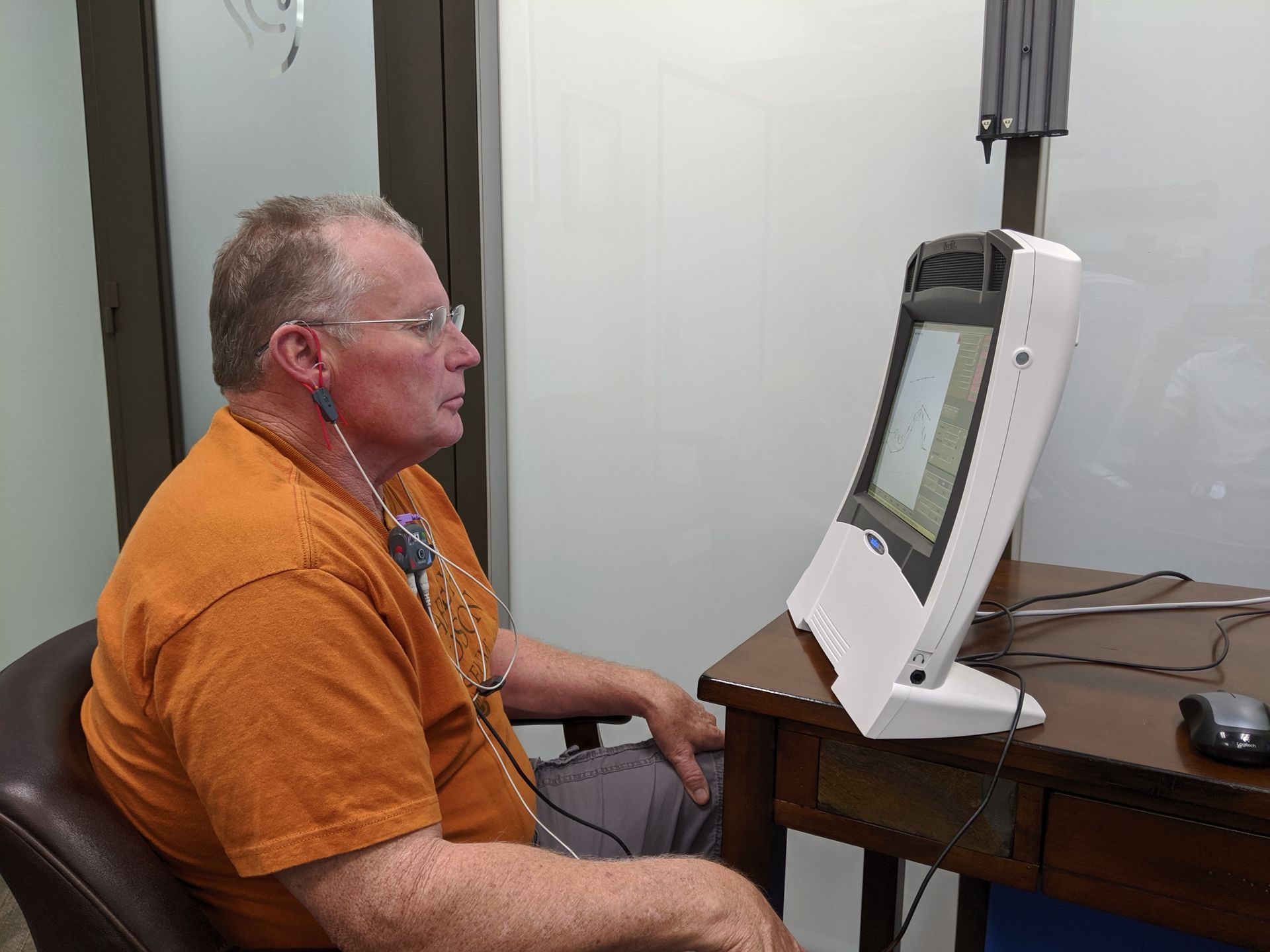 Man in orange shirt undergoes hearing test, looking at a computer screen, in a clinical setting.