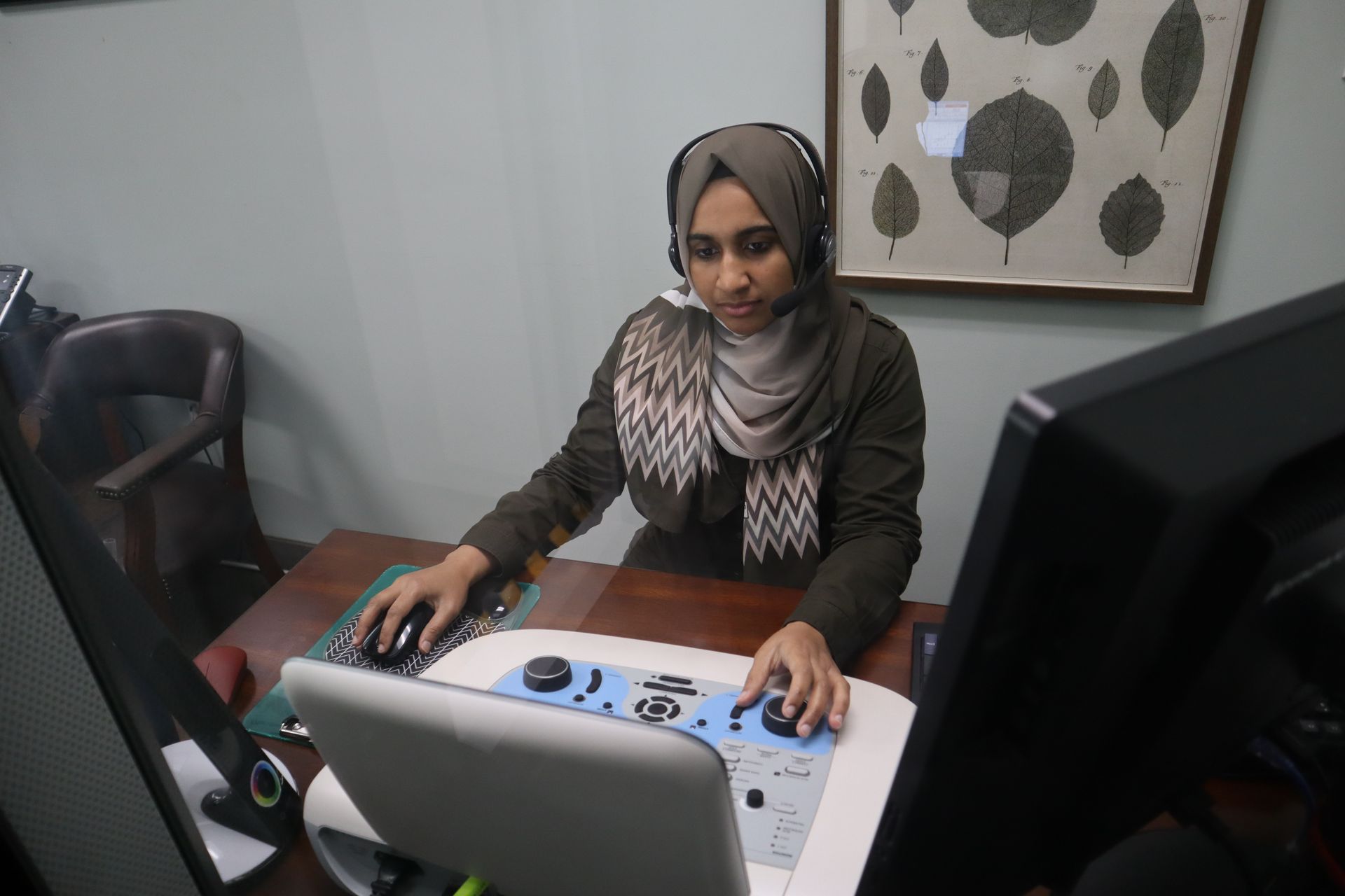 Woman wearing a hijab and headset operates a medical device in an office.