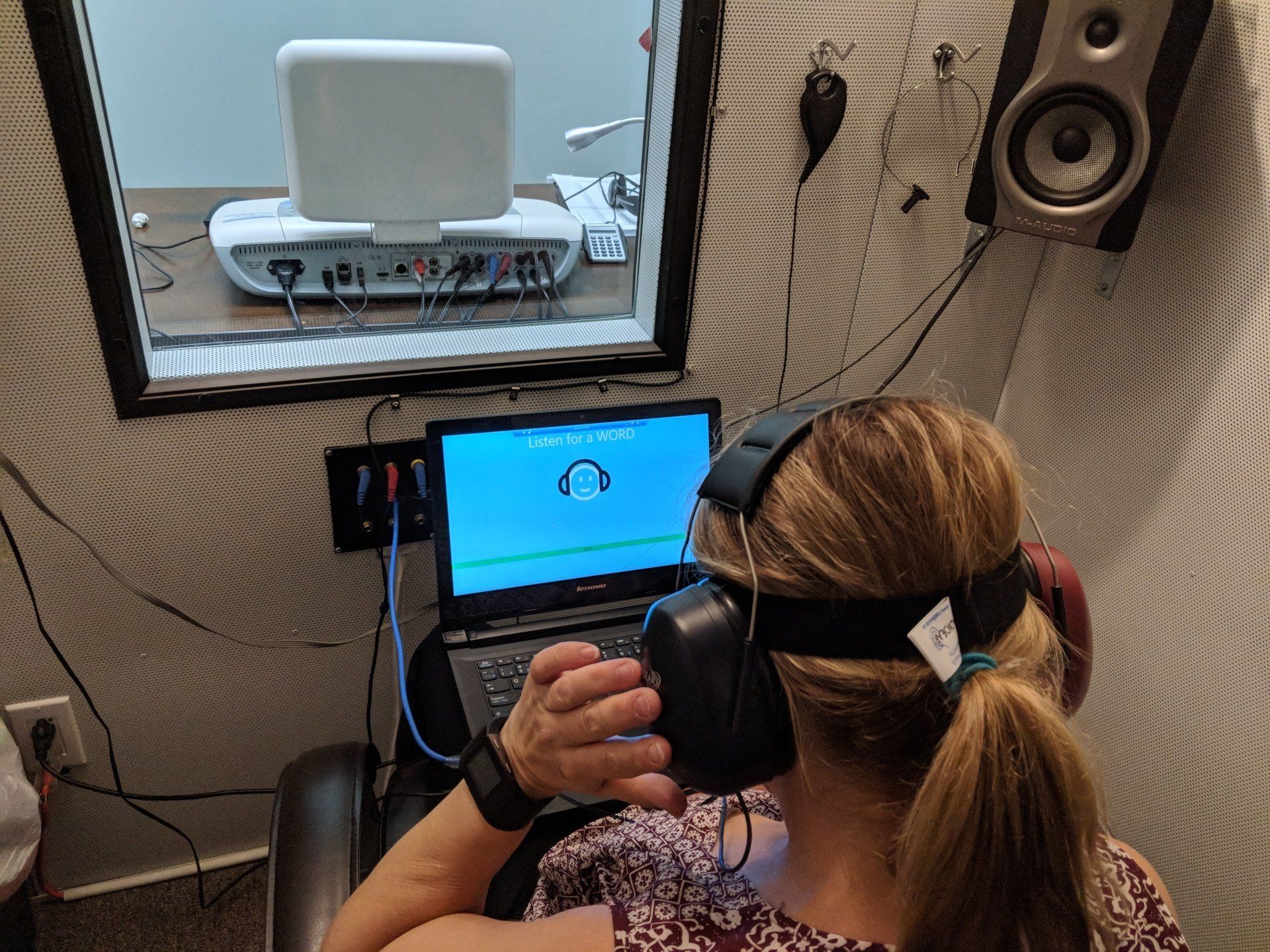 Woman in soundproof booth undergoing hearing test, wearing headphones, looking at laptop.