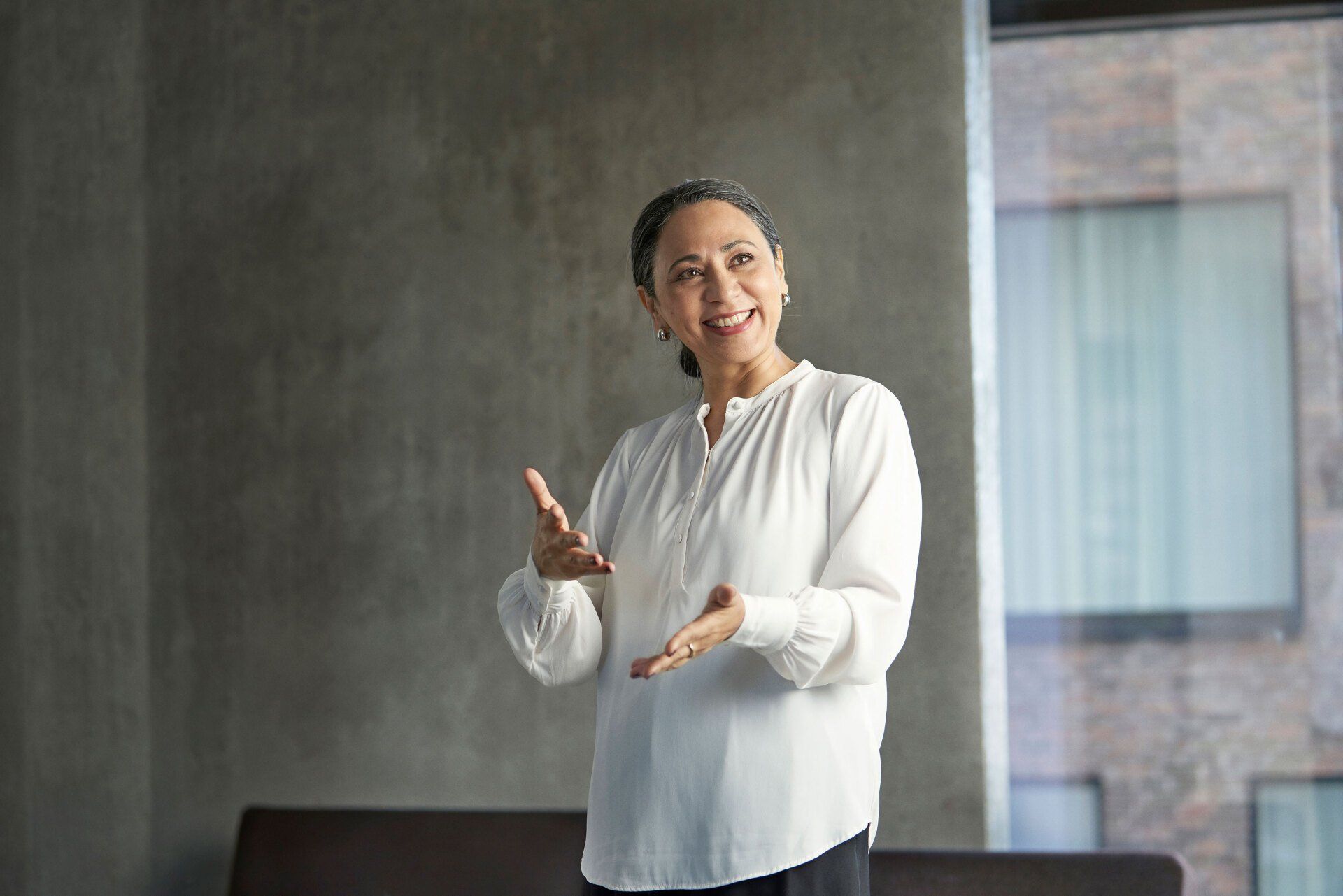 A woman in a white shirt is standing in front of a window and smiling.